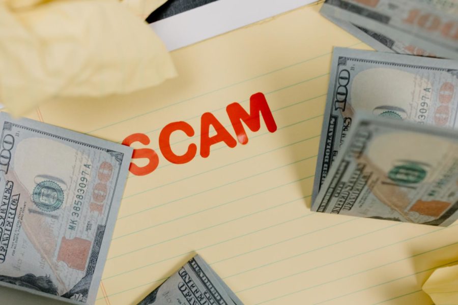 A photo illustration of a yellow notepad with the word scam written in red bubble letters and several partially folded hundred dollar bills.